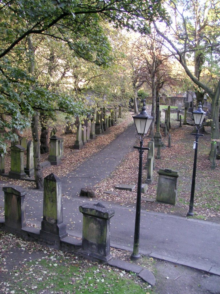 Blick von oben auf einen Friedhof mit vielen laubbedeckten Gräbern und Steingrabsteinen