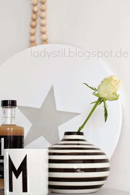 weißer Drehteller mit grauem Stern davor steht eine schwarzweiß getreifte Vase mit einer Rose und ein Designlettersbecher mit einem M