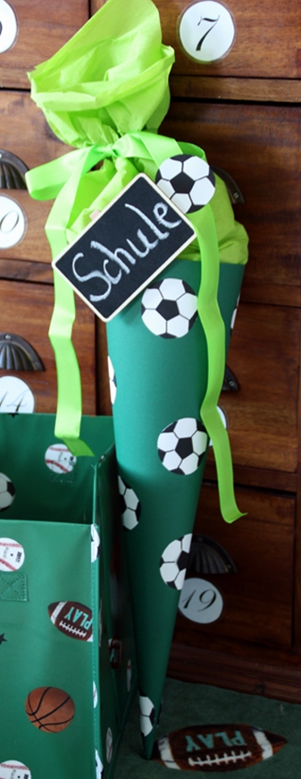 grüne Schultüte mit Fußbällen aus Bällen beklebt und einem kleinen Tafelschild mit Schriftzug "Schule" steht auf einem Badetuch mit Fußballmotiv, daneben eine Aufbewahrungsbox mit Fußballmotiven